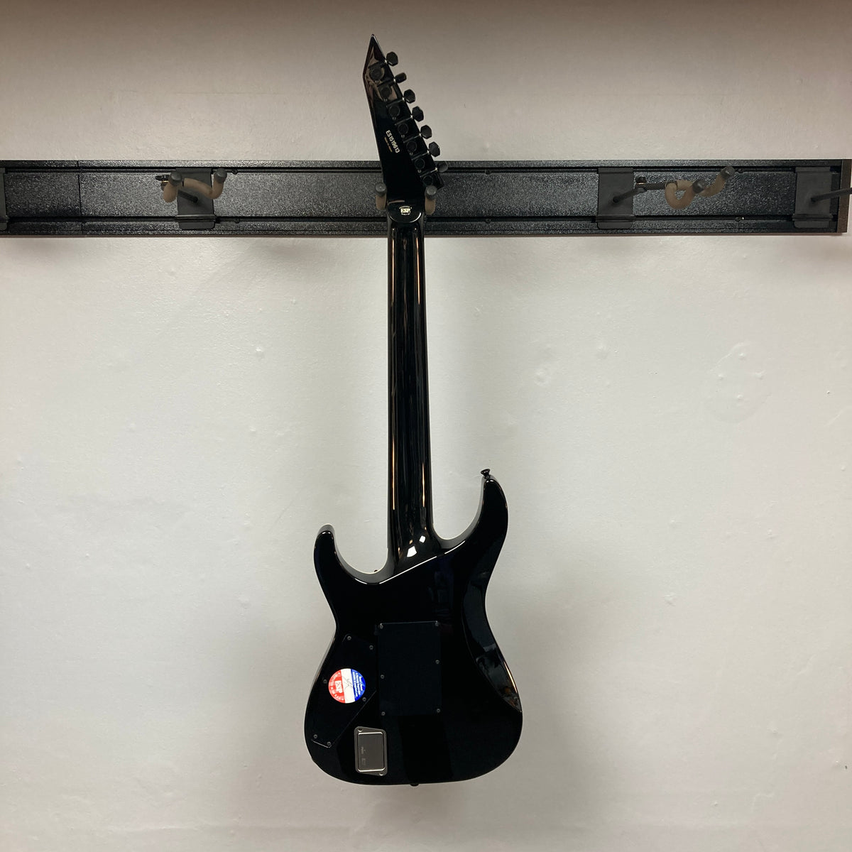 An ESP E-II Horizon FR-7 Black w/Case 7-string electric guitar by ESP hangs on a wall-mounted rack, displaying its back, a sticker near the bottom, and the distinctive Floyd Rose bridge.