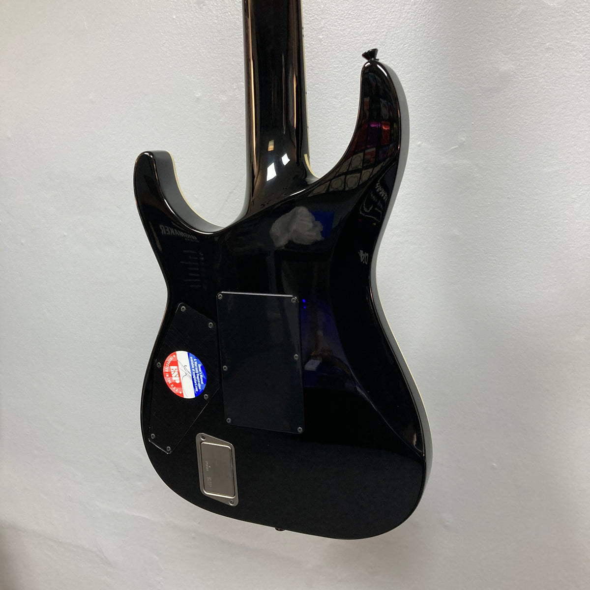 The back of the ESP E-II Horizon FR-7 Black 7-string electric guitar by ESP is shown against a white wall, displaying a sticker and several plastic covers over its electronics.