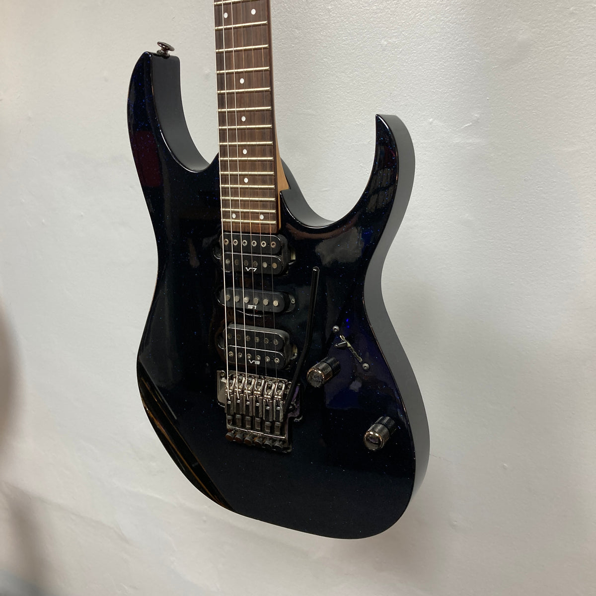 An Ibanez Prestige RG1570 Cosmic Blue electric guitar with a double-cutaway body and tremolo bridge hangs on a white wall.