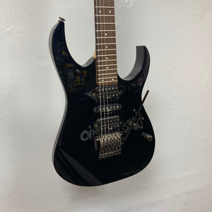 An Ibanez Prestige RG1570 electric guitar in Cosmic Blue with a case, featuring a double-cutaway body, three pickups, and a Wizard Prestige neck, hangs on a white wall.