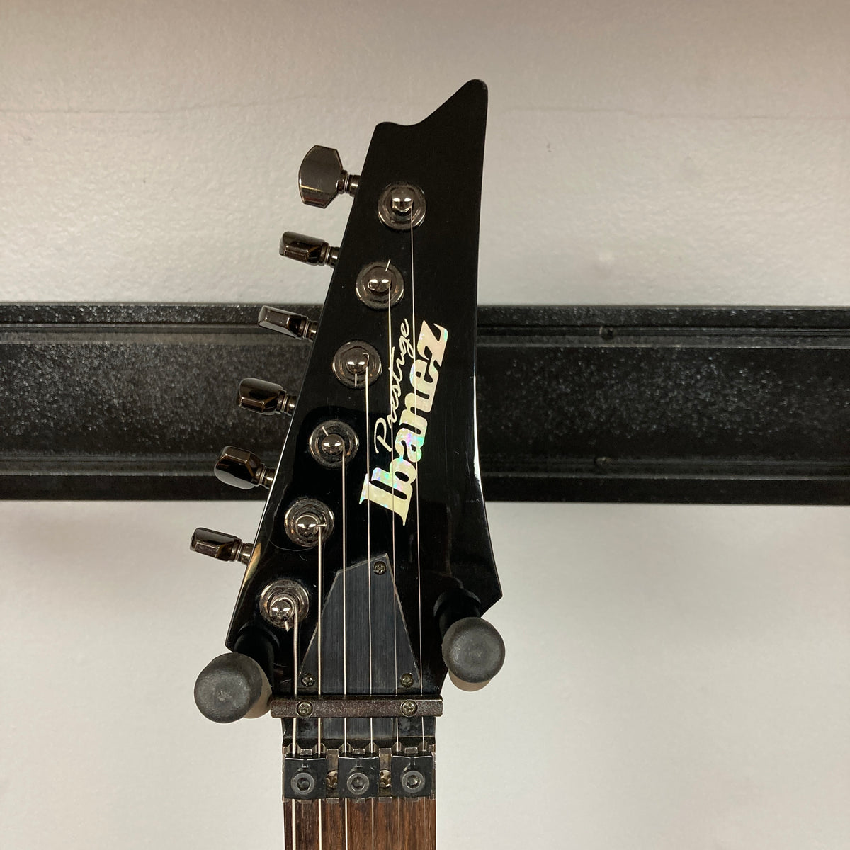 Close-up of the Ibanez Prestige RG1570 Cosmic Blue electric guitar headstock, featuring tuning pegs, logo, and its slim Wizard Prestige neck, shown against a plain wall background. Includes case.