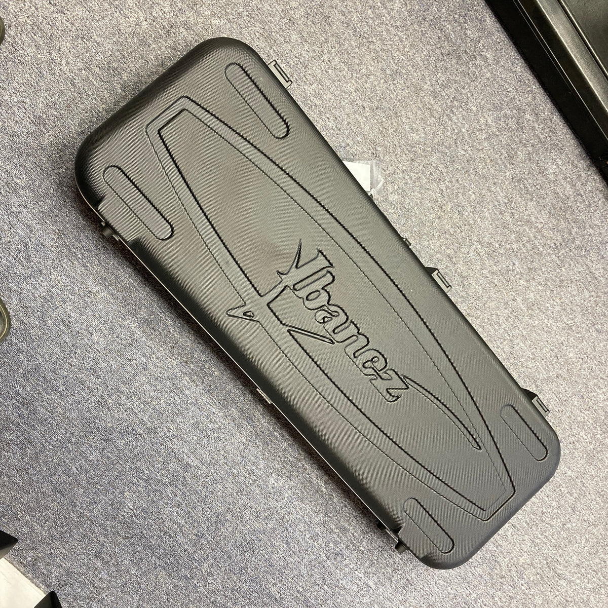An Ibanez Prestige RG1570 Cosmic Blue guitar with a black hard case featuring the Ibanez logo, ideal for this Japanese-made model, is shown resting on a gray carpeted floor.