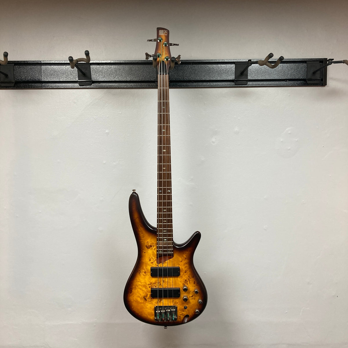 An Ibanez SDGR SR500PB four-string electric bass guitar by Ibanez, featuring a sunburst finish and flatwound strings, hangs on a wall rack against a plain white background. Includes hard case.