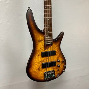 A close-up of the Ibanez SDGR SR500PB bass guitar by Ibanez, featuring a wood grain finish and multiple control knobs, displayed with its hard case against a plain white wall.