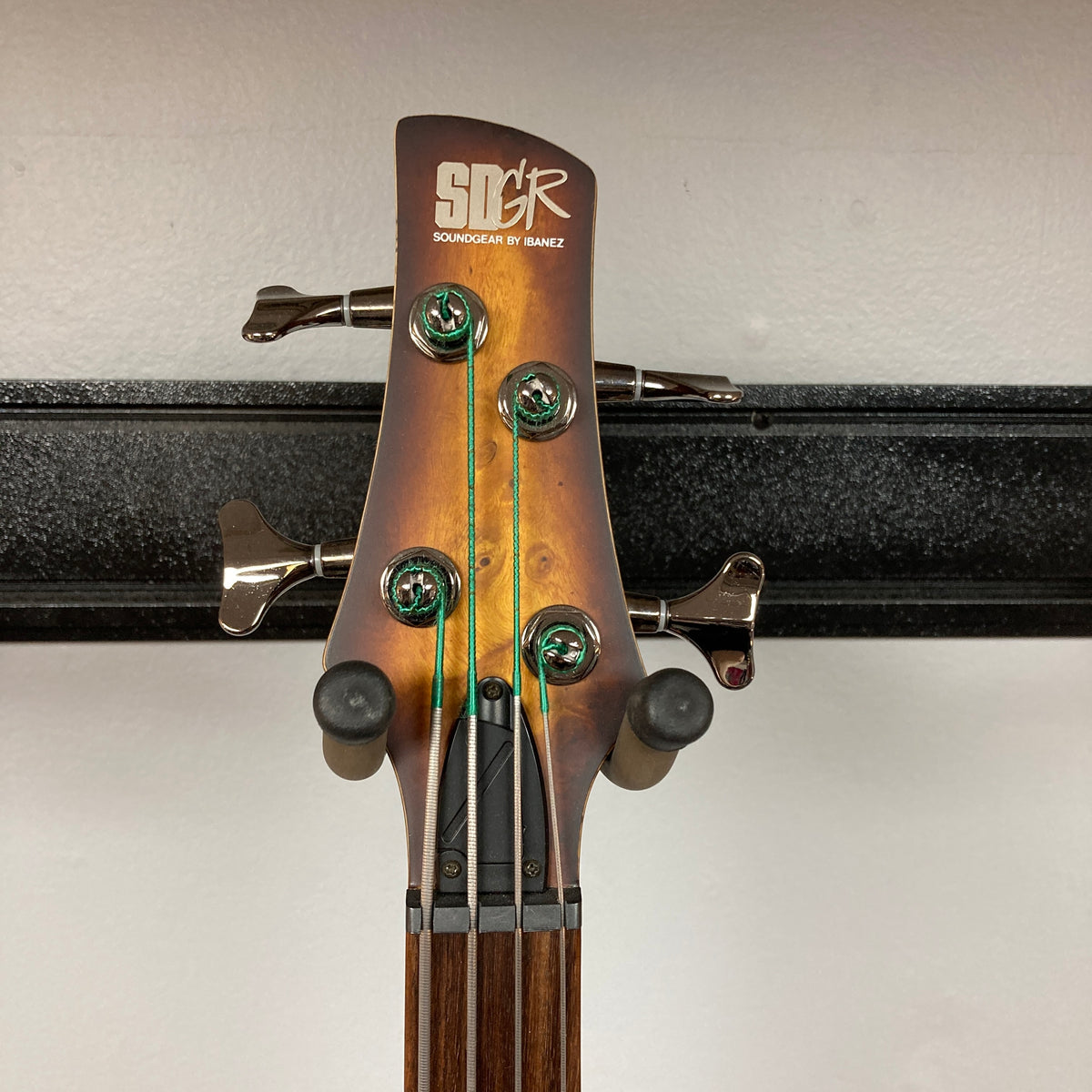 Headstock of an Ibanez SDGR SR500PB bass guitar with four tuning pegs and green flatwound strings, mounted on a wall hanger. Brand: Ibanez. Model: SDGR SR500PB w/Hard case.