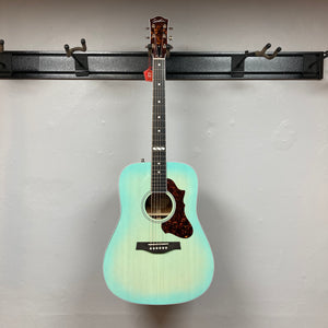 A Godin Imperial Laguna Blue GT EQ SF Acoustic Guitar w/Gigbag hangs on a wall-mounted holder against a plain white wall.