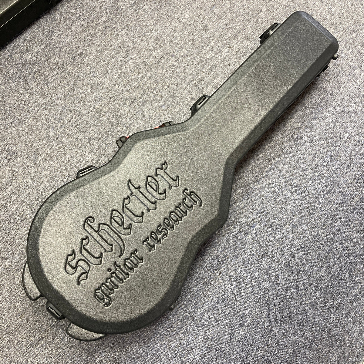 A Schecter Solo-II Custom Trans Black Burst with case, branded "Schecter," lies on a gray carpeted floor. The case is hard, black, and embossed with "Schecter guitar research," made to fit the Solo-II Custom model.