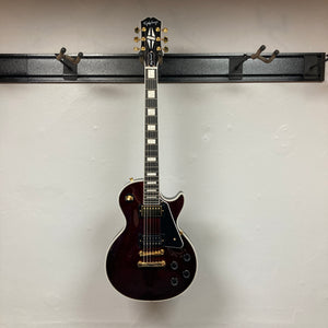 An Epiphone Jerry Cantrell Wino Les Paul Custom electric guitar in wine red with gold hardware and white trim hangs on a wall-mounted rack against a plain white background. Comes with case.
