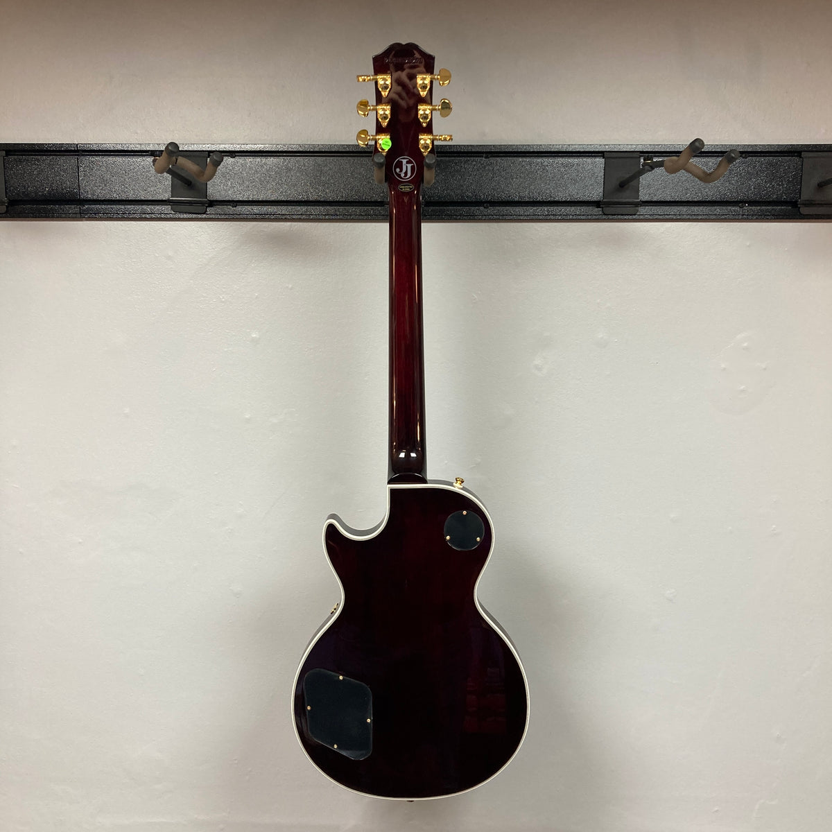 An Epiphone Jerry Cantrell Wino Les Paul Custom in Wine Red with gold tuning pegs and white trim, shown from the back, hangs on a wall-mounted rack. Case included.