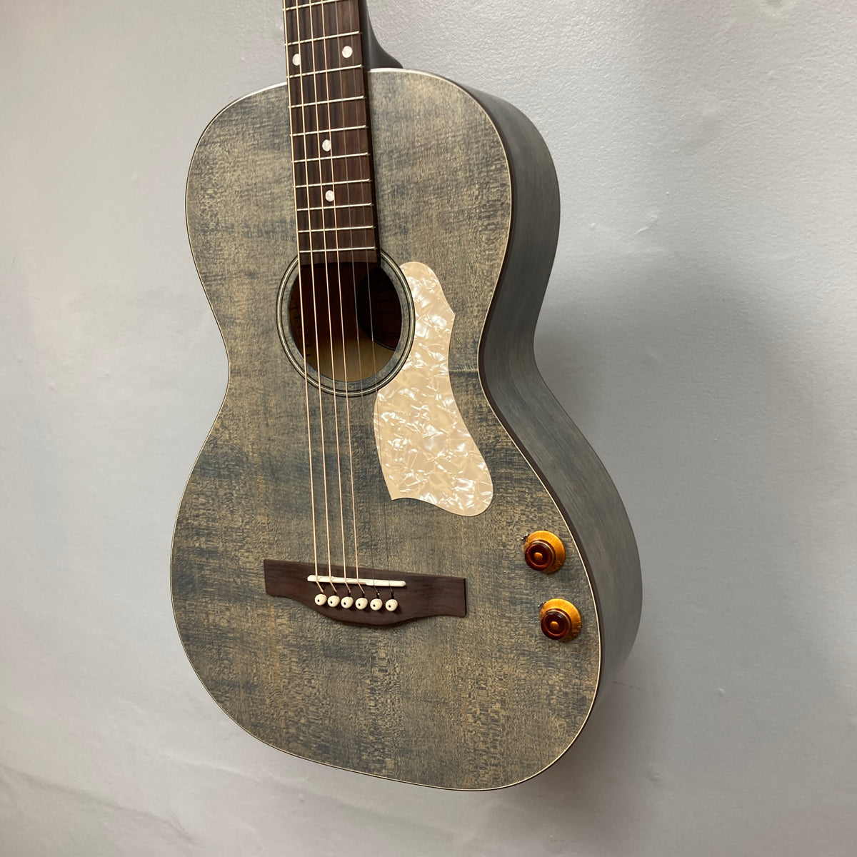 An Art & Lutherie Roadhouse Denim Blue Q-Discrete acoustic guitar with six strings, two control knobs, a white pickguard, and a wooden bridge hangs against a plain light-colored wall.
