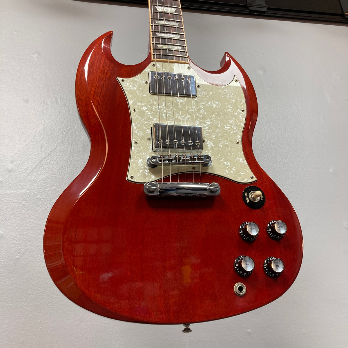 A Gibson SG Standard Heritage Cherry electric guitar (year 2000, Consign) is hanging on the wall, featuring a white pickguard and four control knobs.