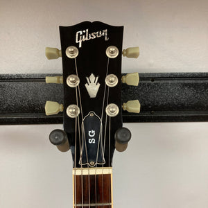 Close-up of the headstock on a Gibson SG Standard Heritage Cherry yr 2000 Consign, showing tuning pegs, logo, and part of a guitar wall mount.