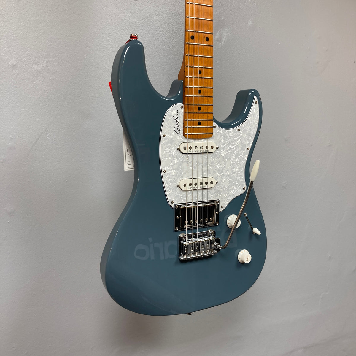 The Godin Session T-Pro Arctik Blue MN SF with Bag is a blue electric guitar featuring a maple neck, white pickguard, Seymour Duncan HSS pickups, tremolo bridge, and control knobs. Displayed against a plain gray wall.