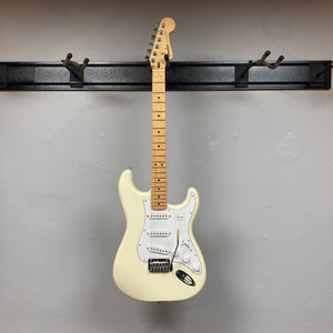 A Fender Standard Stratocaster Olympic White Refurb guitar by Fender hangs on a black wall-mounted rack against a light gray wall.
