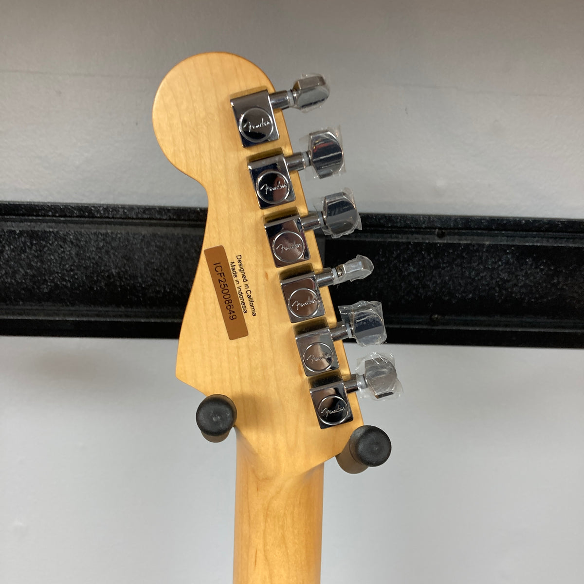 Back of a Fender Standard Stratocaster Olympic White Refurb headstock by Fender, showing six tuning pegs, serial number, and manufacturer info, resting on a wall-mounted guitar holder.