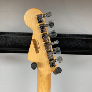 Back of a Fender Standard Stratocaster Olympic White Refurb headstock by Fender, showing six tuning pegs, serial number, and manufacturer info, resting on a wall-mounted guitar holder.