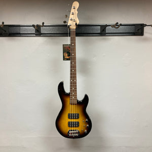 A G&L L-2000 USA Tobacco Sunburst Bass w/Case (used) with four strings hangs on a wall-mounted rack against a plain white wall.