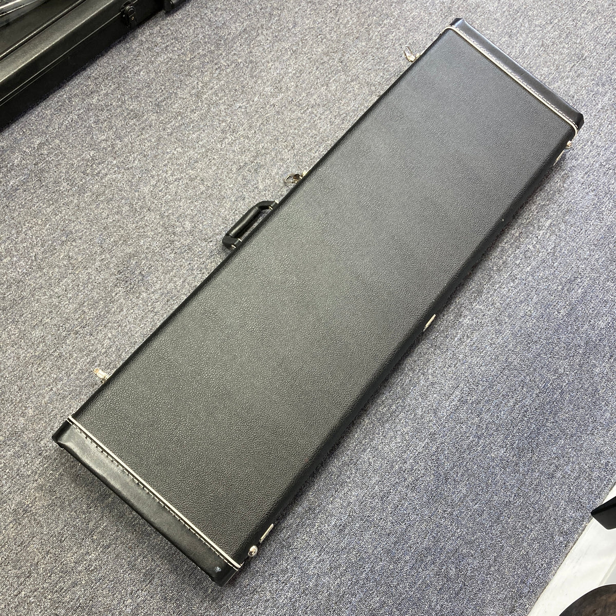 A closed, rectangular black case with a handle—likely containing a used G&L L-2000 USA Tobacco Sunburst Bass—rests on a gray carpeted floor.