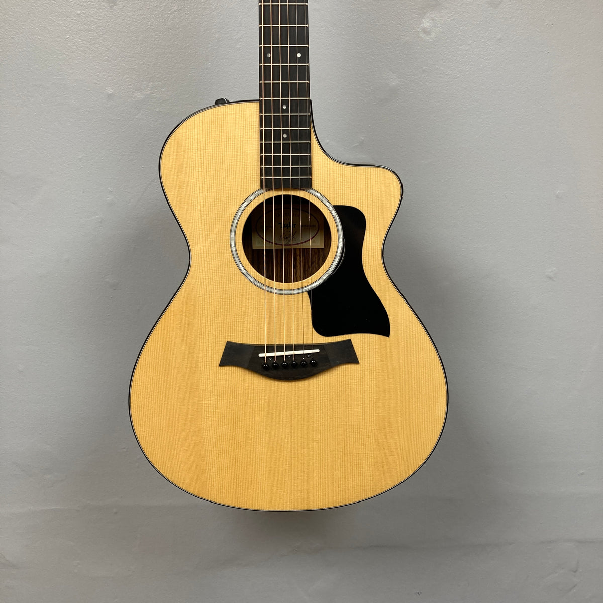 A used Taylor 212ce Plus w/Gigbag acoustic guitar by Taylor, with a natural finish and black pickguard, is shown against a plain light gray wall.