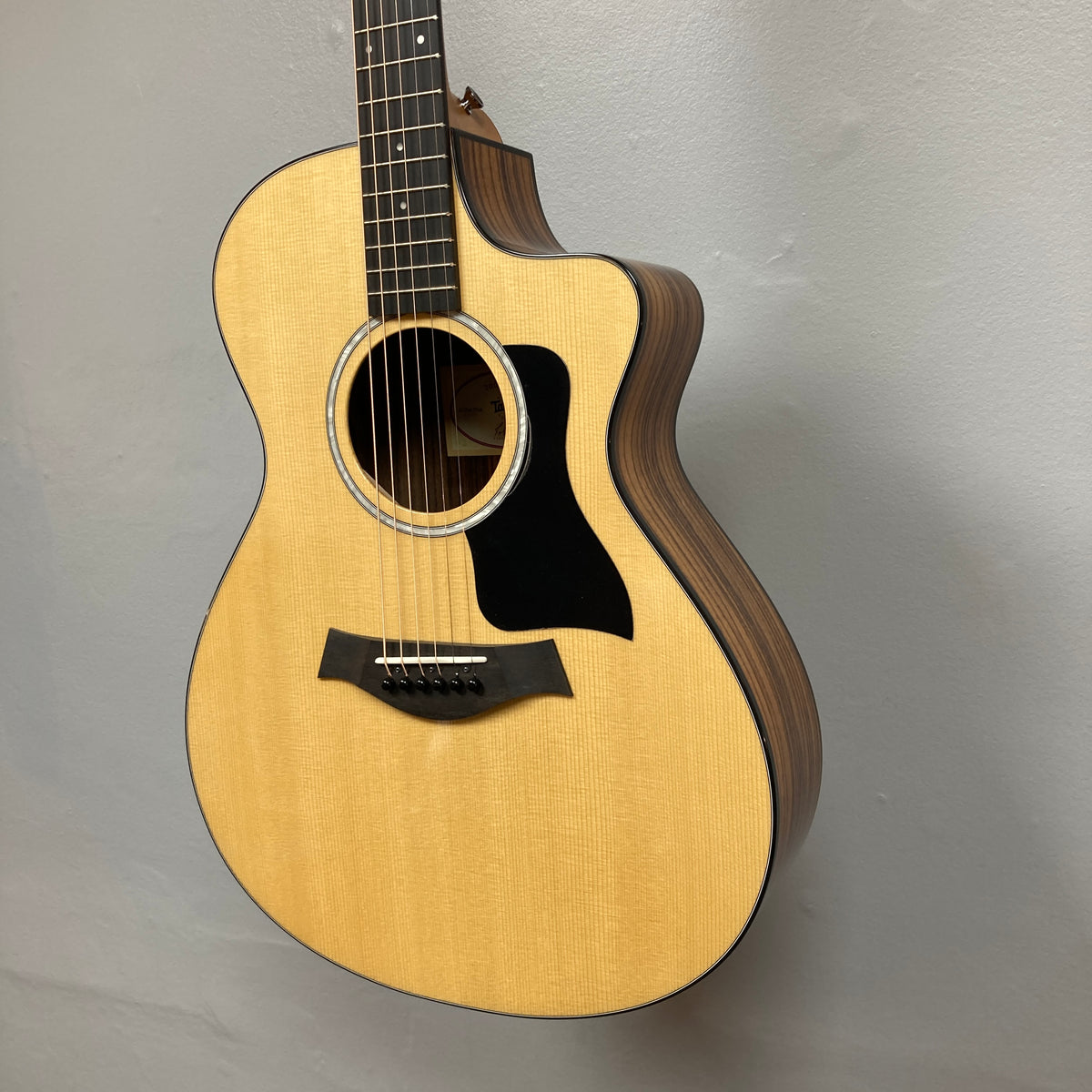 Taylor 212ce Plus w/Gigbag Used Consign by Taylor, featuring a light wood top, dark wood sides, and black pickguard, is displayed hanging on a light gray wall.