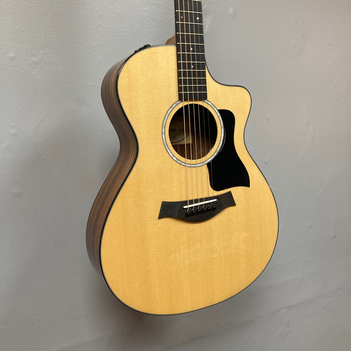 Taylor 212ce Plus w/Gigbag Used Consign by Taylor, featuring a light wood top, cutaway body, and dark wood sides, displayed on a light gray wall.