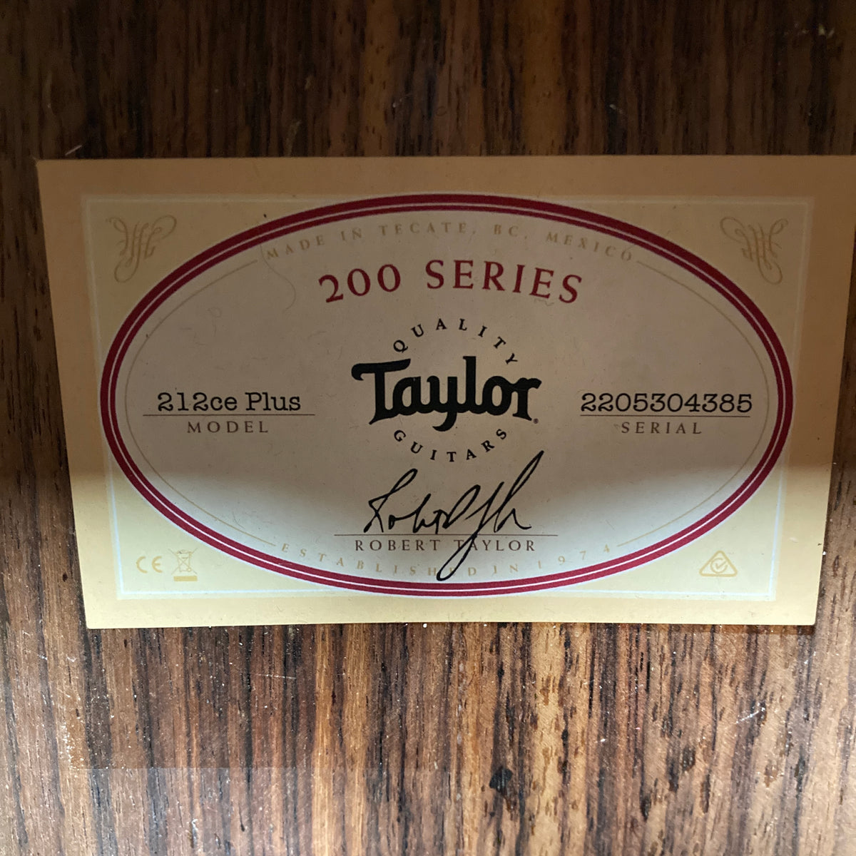 Taylor 212ce Plus w/Gigbag Used Consign label features the model name and signature, highlighting Taylor’s renowned craftsmanship on a wooden surface—quality from the makers of iconic acoustic guitars.