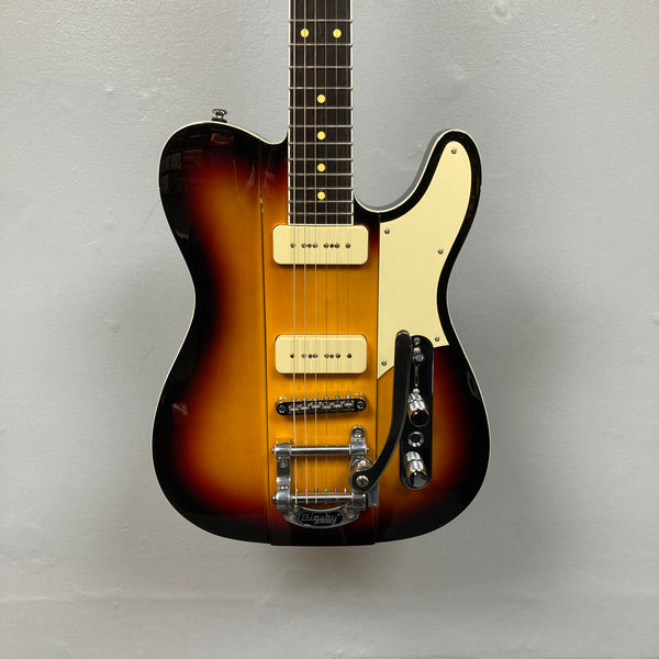 The Reverend Greg Koch Signature Gristle 90 3-Tone Burst electric guitar features two cream Fishman pickups, a white pickguard, and a Bigsby vibrato tailpiece, shown here against a plain gray wall.