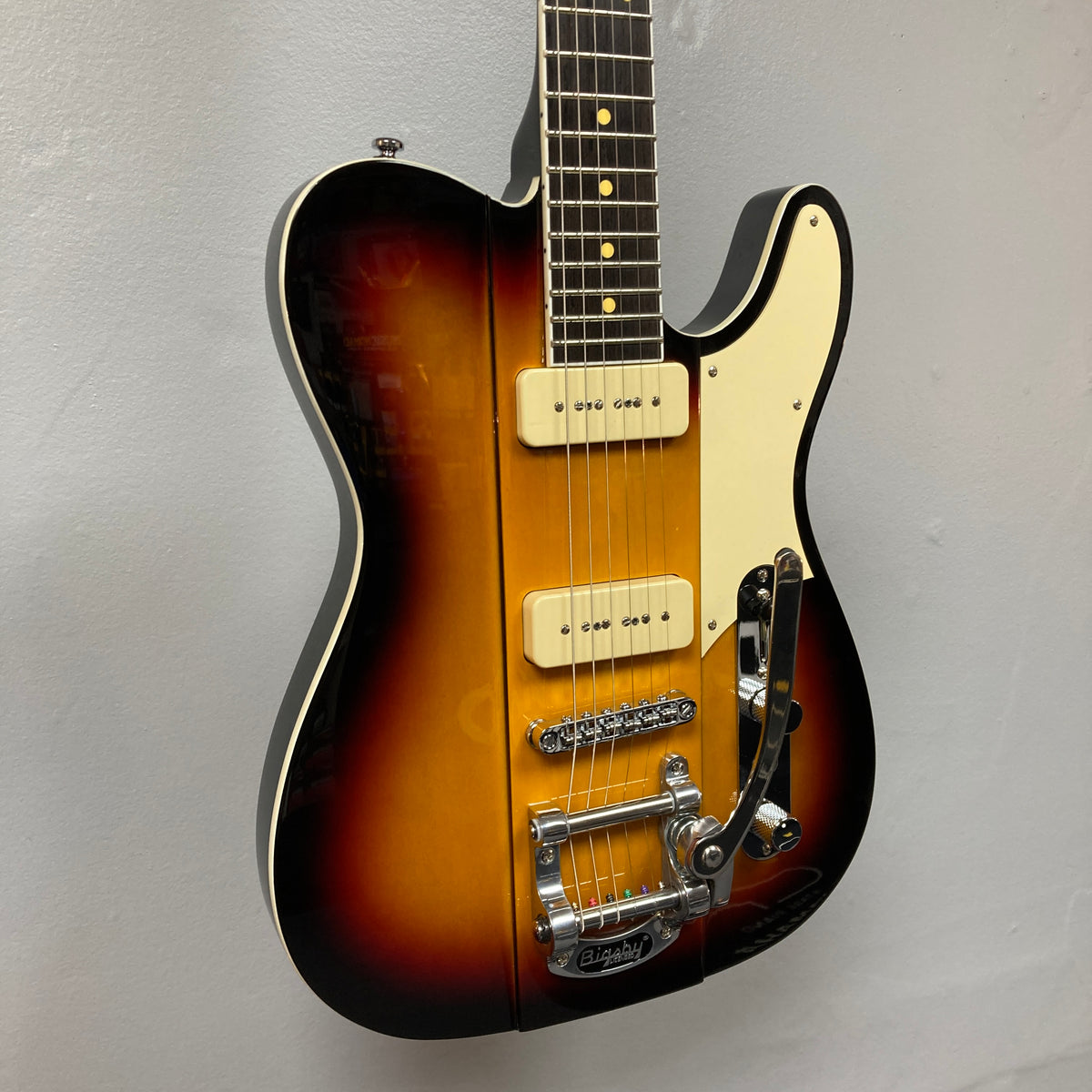 A Reverend Greg Koch Signature Gristle 90 electric guitar in 3-Tone Burst with a white pickguard, two cream-colored Fishman Gristle 90 pickups, Bigsby tremolo, and chrome hardware is shown against a plain gray wall. Brand: Reverend.