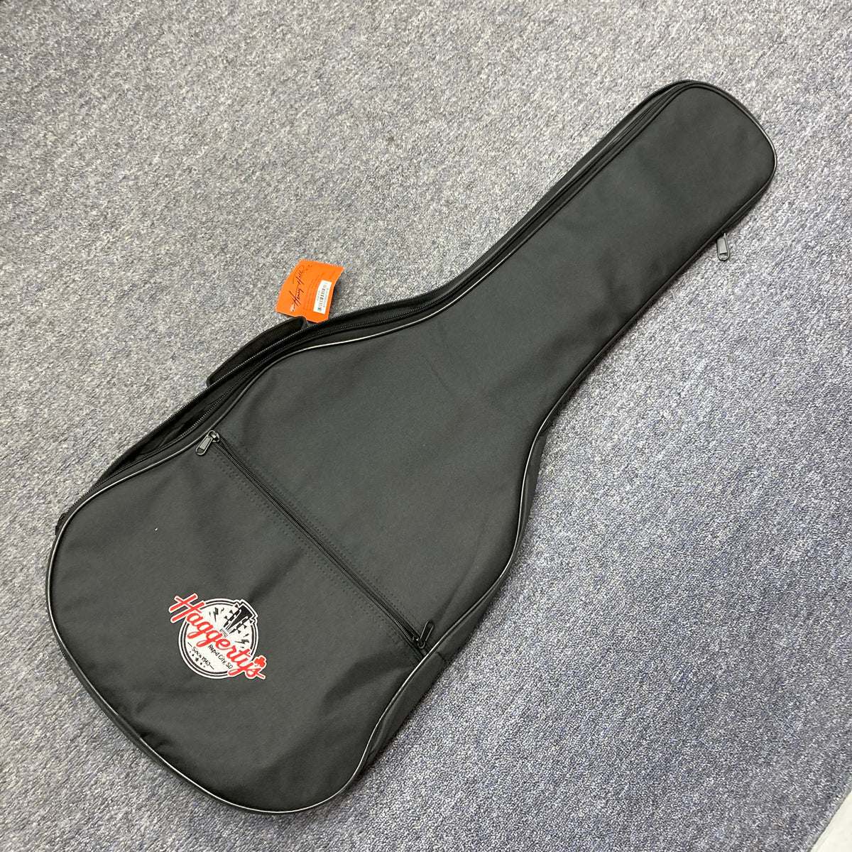 A black soft gigbag with a front zippered pocket and a "Haggleco" logo, perfect for your Reverend Greg Koch Signature Gristle 90 3-Tone Burst electric guitar, rests on a gray carpeted floor.