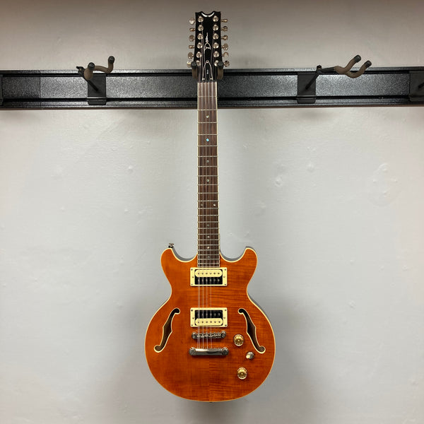 The Dean Boca Trans Amber Burst 12 String w/Gigbag by Dean is shown with a striking amber burst finish, twelve strings, two pickups, four control knobs, and two f-holes against a plain light gray background.