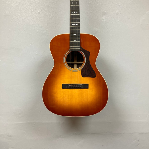 The Guild Gad-30reasb w/Case, an orchestra-size acoustic-electric guitar with a sunburst finish, is hanging on a plain, light-colored wall and features a Fishman Matrix pickup for exceptional amplified sound.