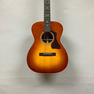 The Guild Gad-30reasb w/Case, an orchestra-size acoustic-electric guitar with a sunburst finish, is hanging on a plain, light-colored wall and features a Fishman Matrix pickup for exceptional amplified sound.