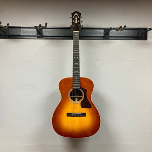 A Guild Gad-30reasb w/Case, an orchestra-size acoustic-electric guitar with a striking sunburst finish, hangs on a black wall-mounted rack against a plain white wall. Its solid Indian rosewood back and sides catch the light, while the discreet Fishman Matrix pickup ensures this Guild instrument is both visually stunning and performance-ready.