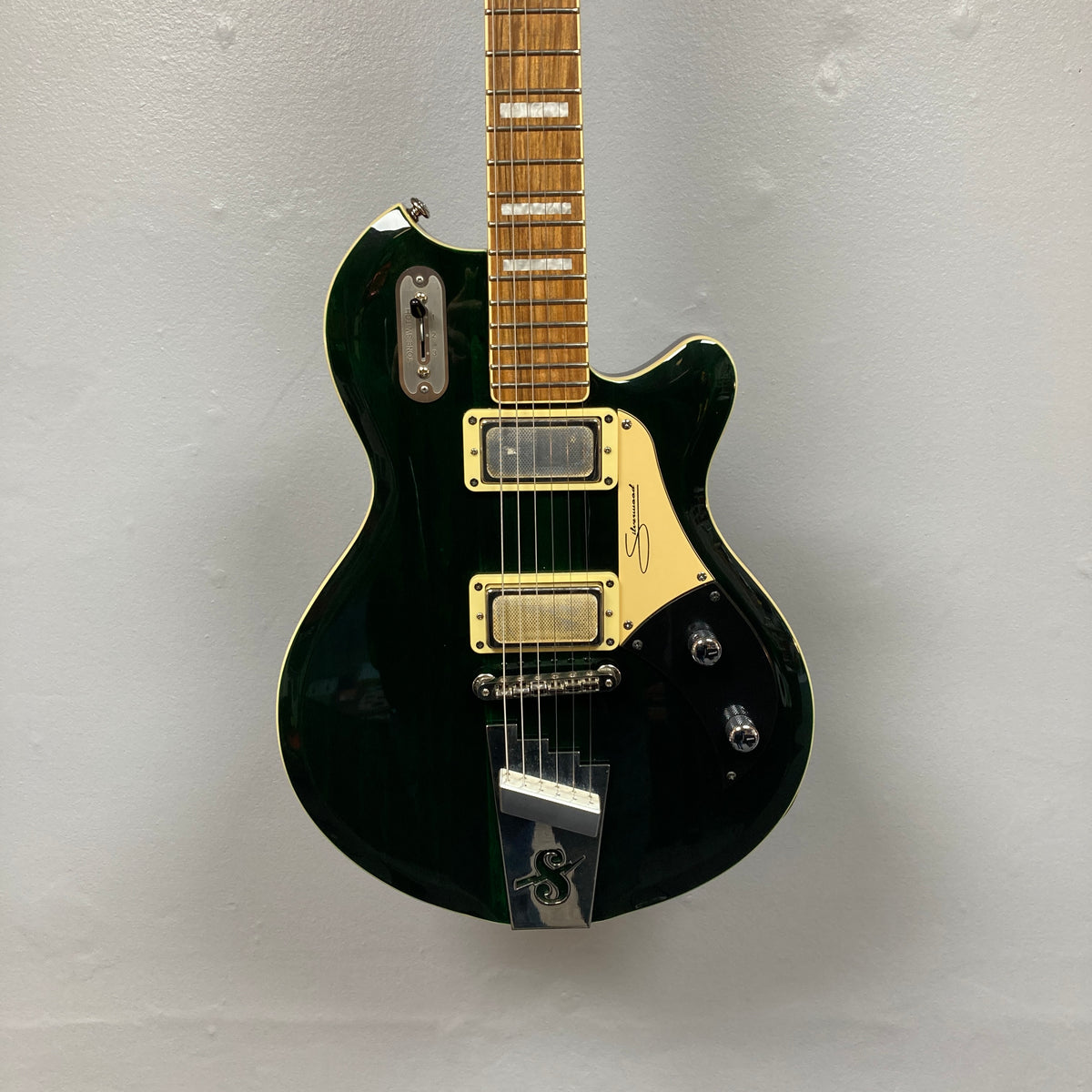 A Supro 1296 Silverwood in British Racing Green, featuring two Gold Foil pickups, a gold pickguard, and chrome hardware, is hanging against a light gray wall.