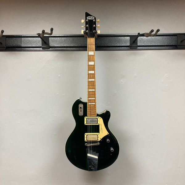 A Supro 1296 Silverwood in British Racing Green, featuring two Gold Foil pickups, a gold pickguard, and chrome hardware, is hanging against a light gray wall.
