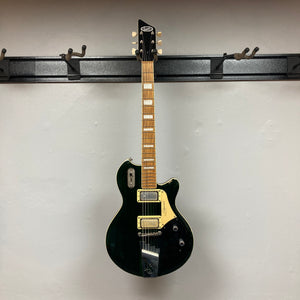 A Supro 1296 Silverwood in British Racing Green, featuring gold hardware and unique Gold Foil pickups, hangs on a wall-mounted rack. The eye-catching finish of this electric guitar stands out beautifully against the white background.