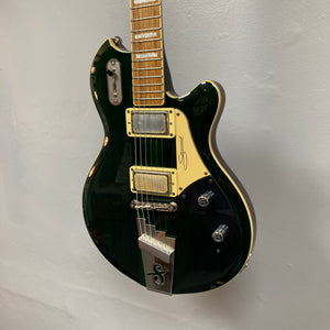 Supro 1296 Silverwood electric guitar in British Racing Green with a cream pickguard, two humbucker pickups, and silver hardware—hanging against a plain light gray wall; includes gigbag.