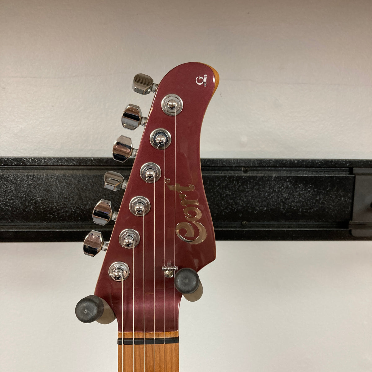 Close-up of the headstock of a Cort FW-G300 PRO Vivid Burgundy electric guitar, featuring six tuning pegs, mounted on a wall hanger against a white background.