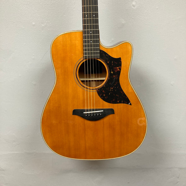 Yamaha A3M ARE Vintage Natural Acoustic Guitar with cutaway body, solid mahogany construction, wood finish, black pickguard, and six strings, pictured against a plain light gray wall.