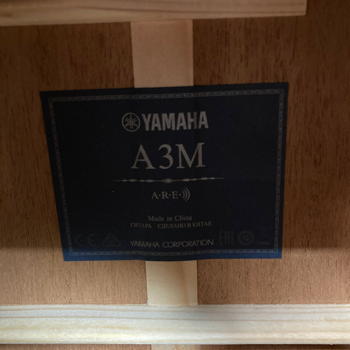 A Yamaha label inside a wooden guitar body displays the model Yamaha A3M ARE Vintage Natural Acoustic Guitar w/Hard Bag Used, indicating it is made in China; it features a solid mahogany build and the advanced SRT2 pickup system.