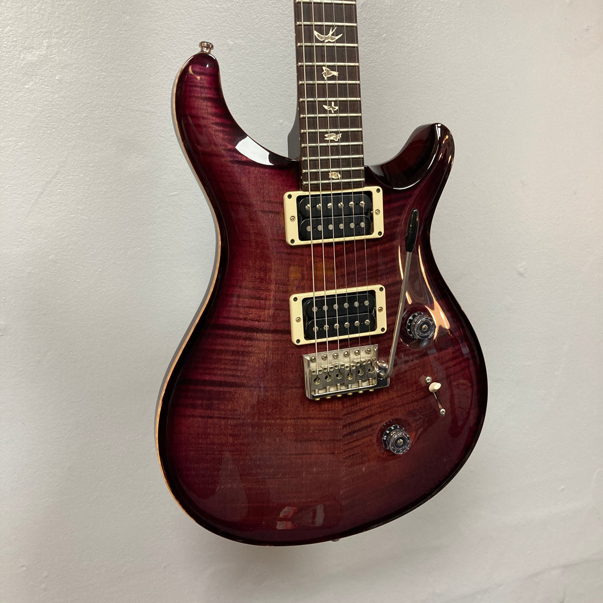 A stunning PRS Custom 24 Violet 2011 electric guitar, featuring a glossy finish, signature bird inlays on the fretboard, two powerful 59/09 humbuckers, and a tremolo bridge, hangs against a plain white wall.