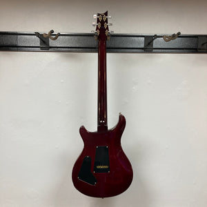 A PRS Custom 24 Violet 2011 electric guitar is shown hanging on a wall mount, photographed from the back against a plain white wall.