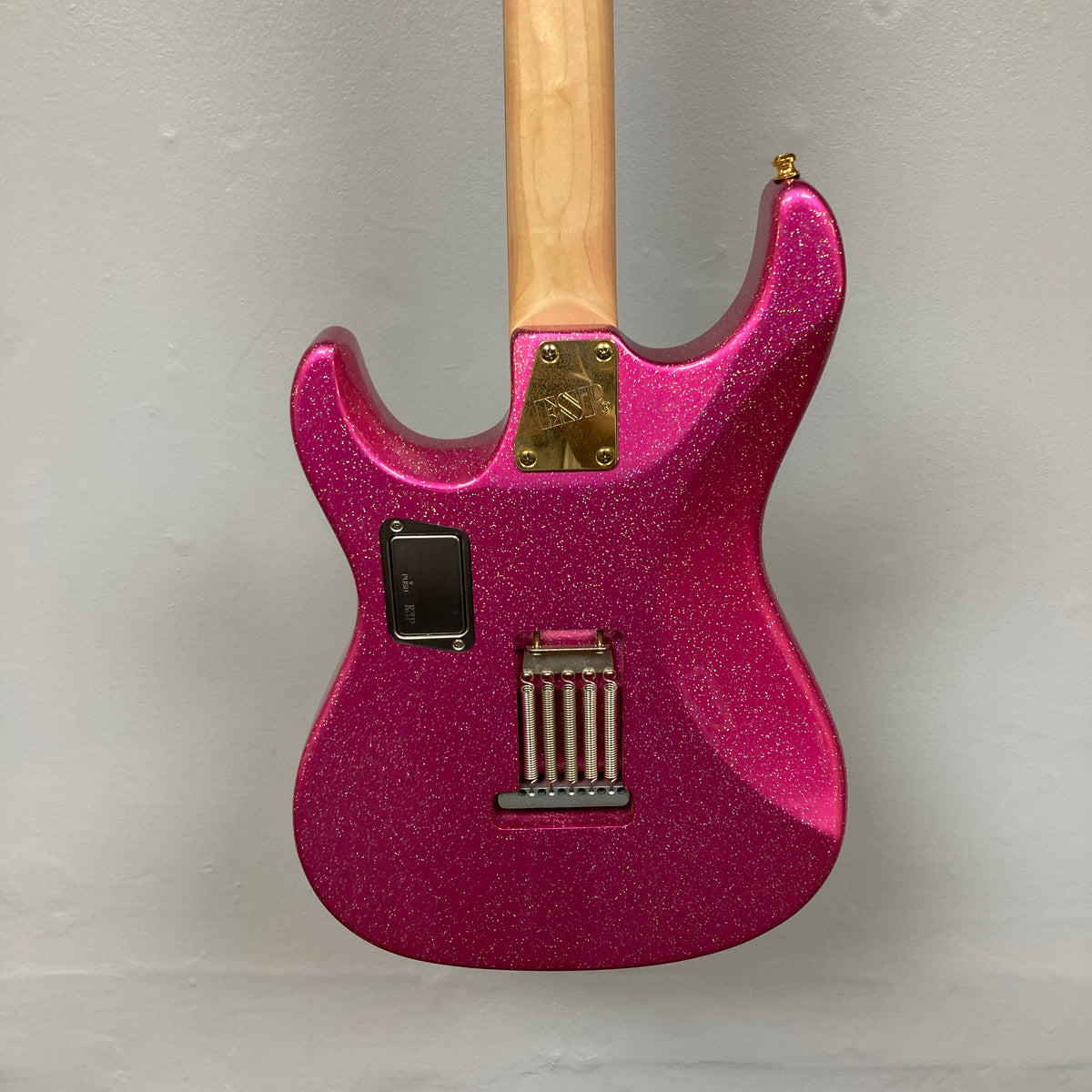 A close-up view of the back of an Edwards ESP E-SN-185-TO Takayoshi Ohmura Twinkle Pink electric guitar, featuring dual humbuckers, a maple neck, metal hardware, and a rectangular battery compartment, set against a plain light gray background.