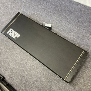A closed black Edwards guitar case with a metal latch sits on a gray carpeted floor, ready to protect your prized Edwards ESP E-SN-185-TO Takayoshi Ohmura Twinkle Pink electric guitar.