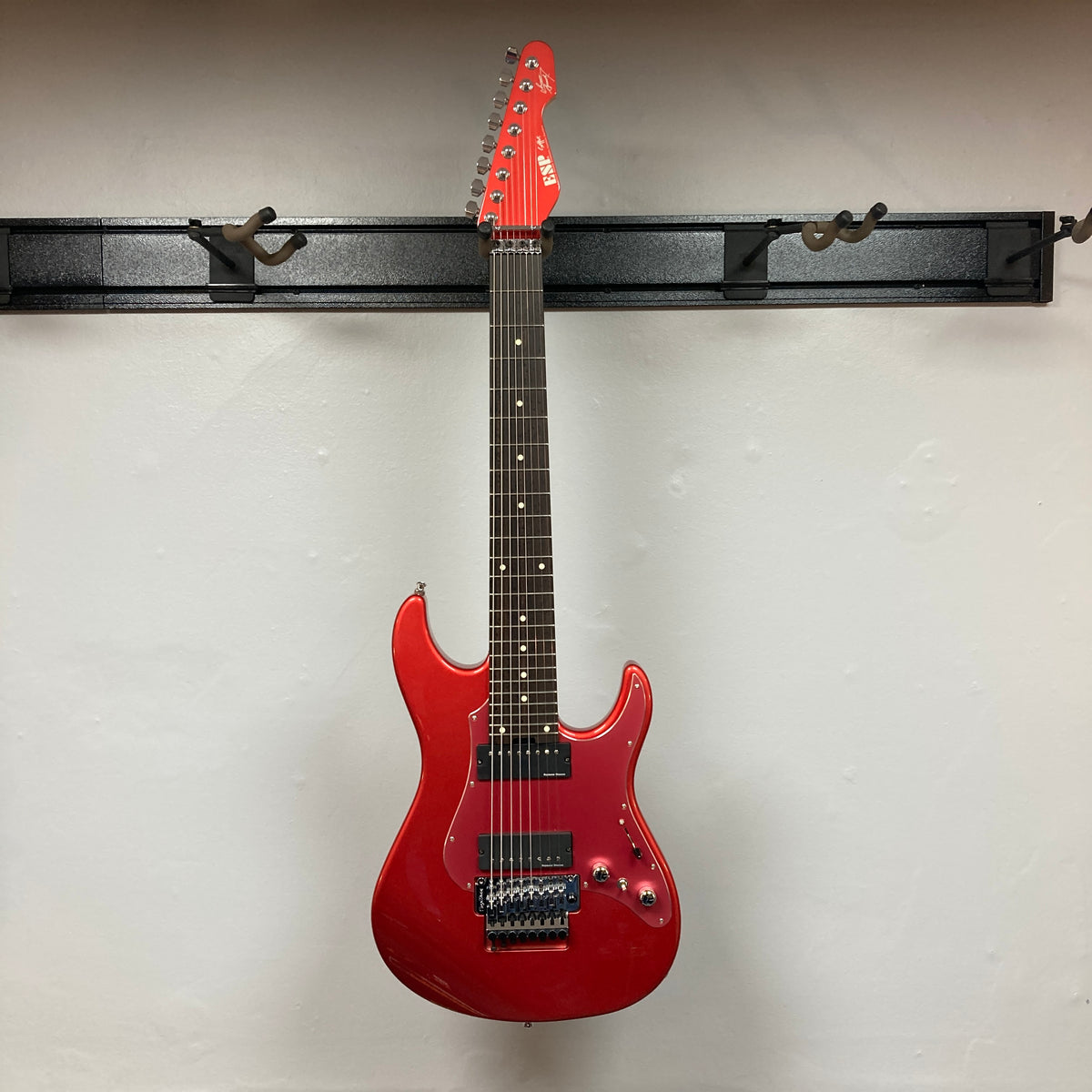Showcased on a wall-mounted guitar hanger against a crisp white backdrop, the ESP Snapper Raiden 8 ISAO Custom w/Gigbag by ESP impresses with its vibrant Raiden Red finish, eight-string configuration, double-cutaway body, and signature Floyd Rose bridge.