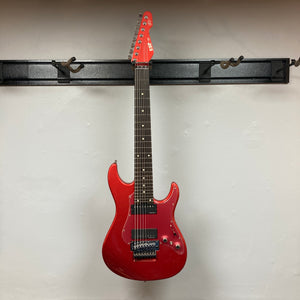 Showcased on a wall-mounted guitar hanger against a crisp white backdrop, the ESP Snapper Raiden 8 ISAO Custom w/Gigbag by ESP impresses with its vibrant Raiden Red finish, eight-string configuration, double-cutaway body, and signature Floyd Rose bridge.