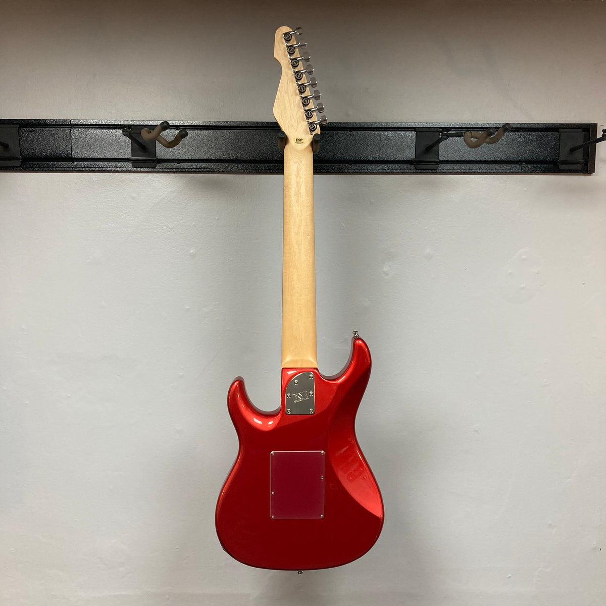 An eye-catching ESP Snapper Raiden 8 ISAO Custom w/Gigbag from ESP is displayed on a black wall-mounted rack, shown from the back with the neck pointing upward against a plain white wall. This remarkable eight-string guitar impresses with its bold Raiden Red finish and contemporary design.