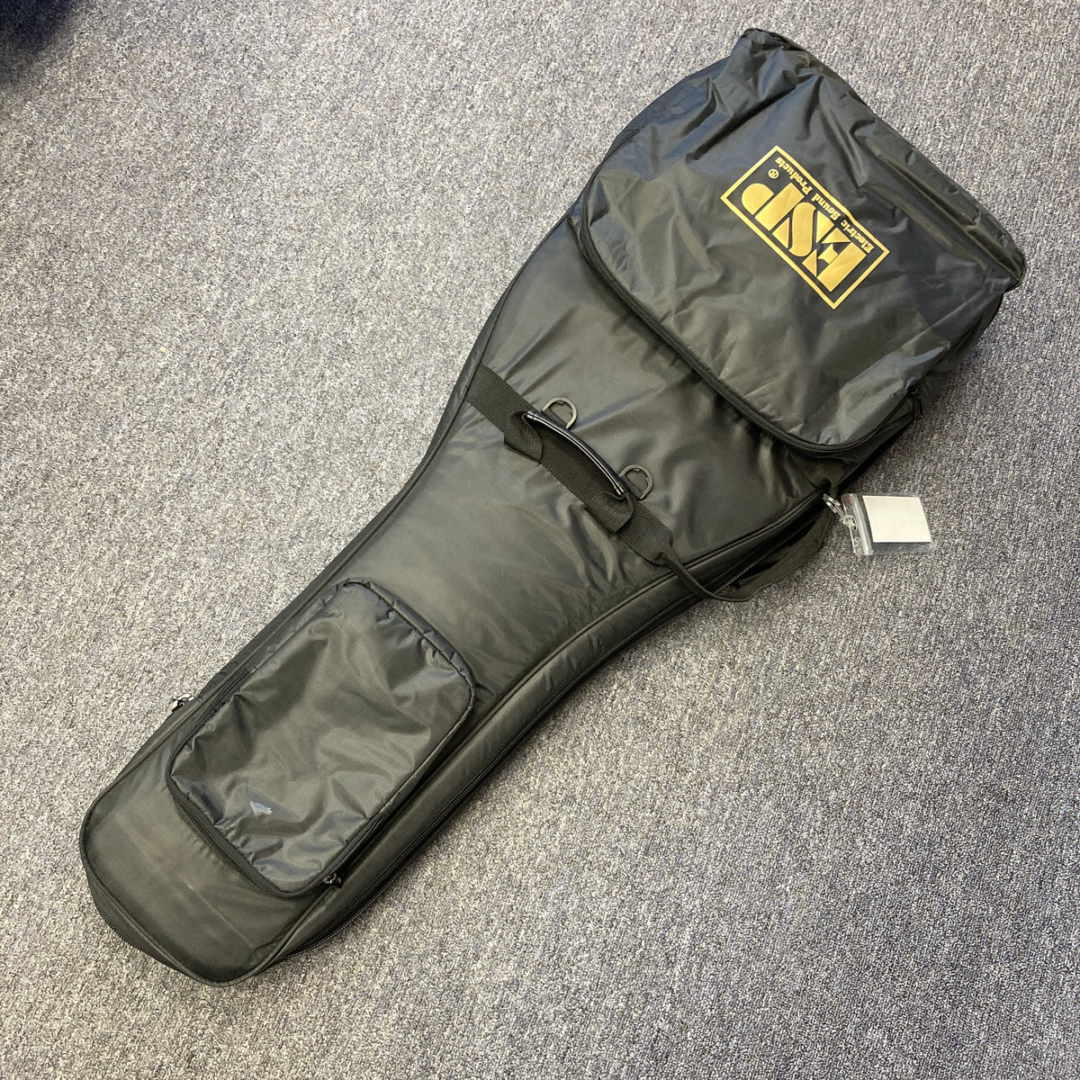 A black padded gig bag, featuring two exterior pockets and a yellow ESP logo, designed for the ESP Snapper Raiden 8 ISAO Custom guitar, rests on a gray carpeted floor.