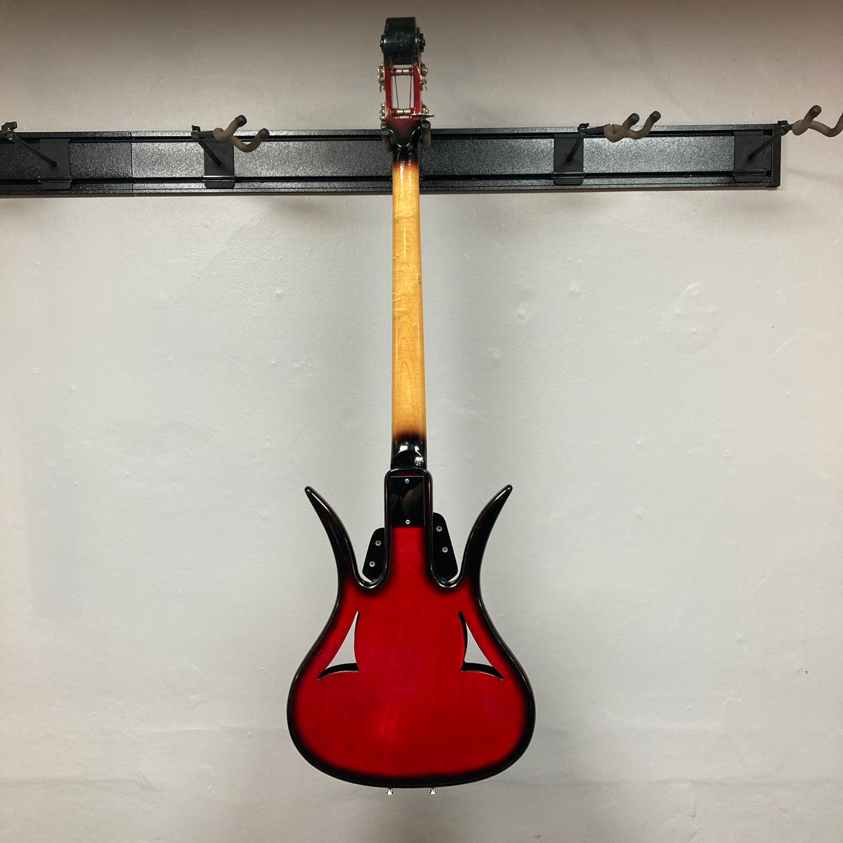 A striking red 1966 Ampeg ASB-1 Devil Bass (Serial #531), featuring a natural wood neck, hangs backward on a black wall-mounted rack to showcase the back of its body and neck, echoing the distinctive vintage design that defines this iconic Ampeg model.