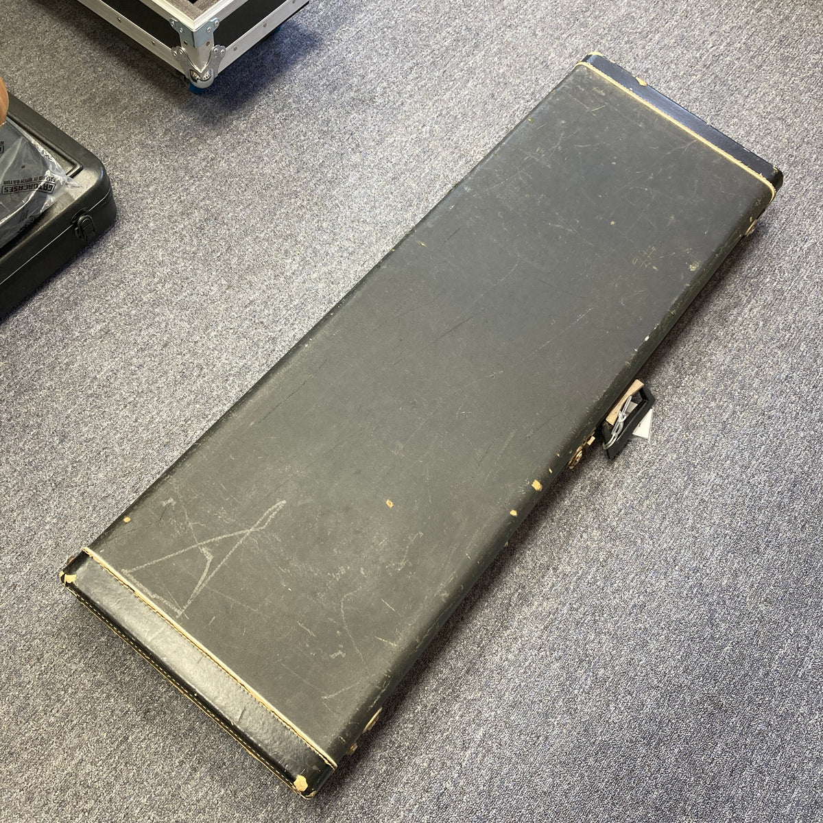 A closed, rectangular black hard case from Ampeg rests on a blue carpeted floor. Showing visible scratches and scuffs, the case suggests a history of use, possibly having once protected an instrument like the 1966 Ampeg ASB-1 Devil Bass (Serial #531) with its unique bridge-mounted transducer pickup.